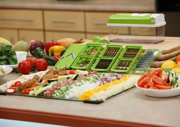 Multifunctional vegetable chopper with stainless steel blades and accessories displayed with sliced vegetables in a kitchen setting.