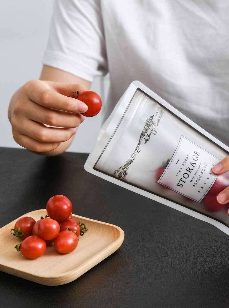 Person placing cherry tomatoes into a reusable silicone ziplock bag with a cup shape, on a black surface.